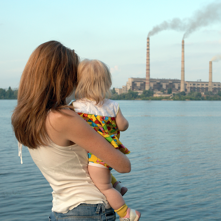 other and child look out across the water at a large industrial plant spewing emissions.