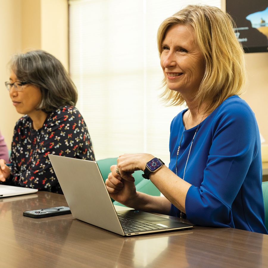 Women sit at a table with open laptops. One woman looks at her screen, while another smiles to someone not pictured.